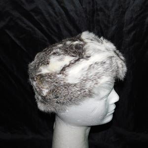 May include: A gray and white fur hat with a soft, fluffy texture. The hat is made of rabbit fur and has a unique, asymmetrical design.