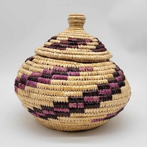 May include: A woven basket with a lid, featuring a natural straw base and purple and brown accents. The basket is round and has a domed lid.