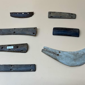 May include: Collection of antique wooden artifacts, including several rectangular pieces with drilled holes, a curved bone-like object, and a dark, curved piece. The items are arranged on a neutral-toned surface.