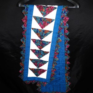 May include: A blue and white quilted table runner with a floral pattern and scalloped edges. The runner is hanging on a black hanger.