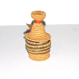 May include: A small, woven basket with a lid, in natural straw tones, with green and purple stripes around the base. The lid has a small, round straw knob and a red band. A small tag is attached.