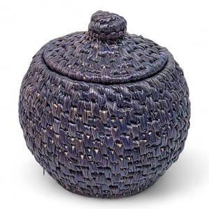 以下が含まれることがあります： 深いインディゴ色の、蓋付きの丸い織りバスケット。バスケットは質感のある表面で、蓋の上部に小さなノブが付いています。バスケットは、おそらく収納や装飾に使用されます。