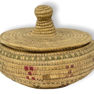 Puede incluir: Una cesta redonda con tapa, tejida con fibras naturales. La cesta presenta una superficie texturizada con tapa y un asa en forma de pomo. La cesta tiene detalles verdes y rojos.
