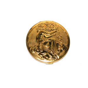 Peut inclure: Un poudrier rond en métal doré avec un motif en relief sur le couvercle. Le design central représente une personne assise entourée de nuages stylisés et d'éléments floraux, le tout sur un fond métallique à la finition texturée.