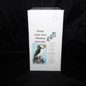 May include: White box with the text "Paint your own Alaskan souvenir." The box features an illustration of a puffin bird and includes a list of contents. The kit contains a ceramic bird statue, paints, and instructions.