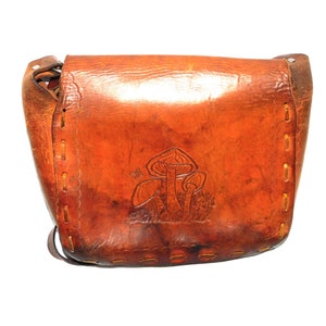 May include: A brown leather satchel bag with a mushroom design embossed on the front. The bag has a flap closure and a long strap.