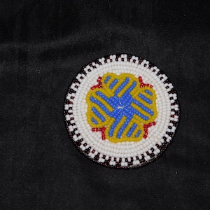 May include: Circular beaded item with a white background and a central design in blue and yellow. The border is white with a dark red and black trim. The item is set against a black background.