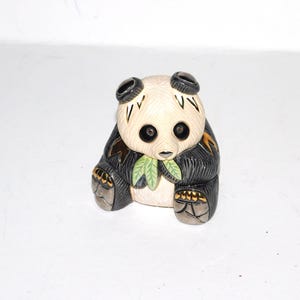 May include: A ceramic panda figurine with black and cream coloring. The panda has black ears, eyes, and paws, with a cream-colored face and body. It holds green leaves and has gold accents.