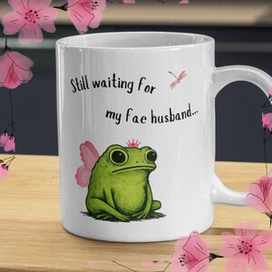 Puede incluir: Taza de cerámica blanca con una rana verde con alas de hada rosas y una corona. La taza tiene el texto "Still waiting for my fae husband..." y una ilustración de libélula rosa. Acentos florales rosas rodean la taza.