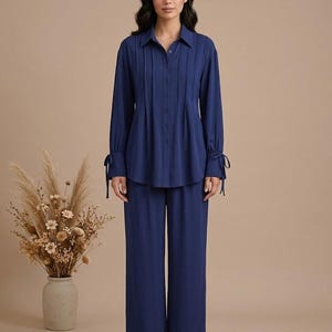 May include: A navy blue two-piece set consisting of a long-sleeved button-down shirt with vertical pleats and tie-cuff details, paired with matching wide-leg trousers. The shirt features a classic collar and a full-length front button closure.