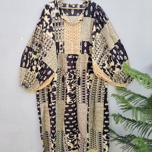 May include: A long, flowing dress with a patchwork design in black, beige, and gold. The dress features a V-neck and wide sleeves, hanging on a wooden hanger against a neutral background. The fabric has a geometric and abstract pattern.