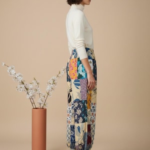 Może przedstawiać: Kobieta w kremowym golfie i spodniach w stylu patchwork z różnymi wzorami kwiatowymi i geometrycznymi. Spodnie mają marszczony kostkę. Białe trampki dopełniają stylizację. Wysoki, terakotowy wazon zawiera białe kwiaty.