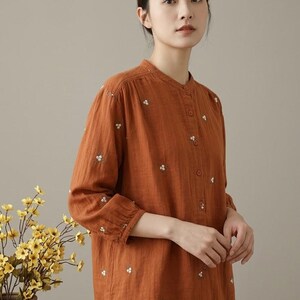 May include: A rust-colored long-sleeve button-down shirt featuring a mandarin collar and small, scattered white floral embroidery. The shirt has gathered sleeve cuffs and a straight hem, paired with blue denim jeans.