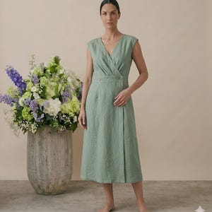 May include: A sage green sleeveless midi dress featuring a V-neckline, a wrap-style bodice, and a fitted waist with two visible buttons. The skirt has a straight silhouette that falls below the knees, crafted from a textured fabric with visible weave.