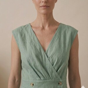 May include: A sage green sleeveless dress featuring a V-neckline with a wrap-style bodice. The garment includes a fitted waistline accented by two round, light brown buttons positioned horizontally on the front of the waist panel.