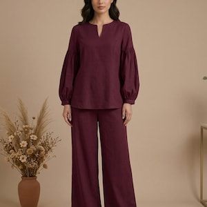 May include: A deep burgundy two-piece set consisting of a long-sleeved blouse with a notched neckline and gathered cuffs, paired with matching wide-leg trousers. The outfit features a solid color and a structured yet relaxed fit throughout the garment.