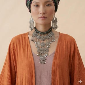 以下が含まれることがあります： 複雑な円形のフィリグリーメダルとぶら下がるチェーンが特徴のシルバー調のステートメントネックレスと、お揃いの長いシャンデリアイヤリング。ジュエリーセットは、モーブ色のトップスとテクスチャーのあるオレンジ色のオープンフロントカーディガンの上に着用されています。