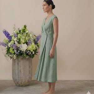 May include: A sage green sleeveless wrap dress featuring a V-neckline, a fitted bodice with a single button closure at the waist, and a midi-length skirt that falls in a straight silhouette. The garment is made of a textured fabric.