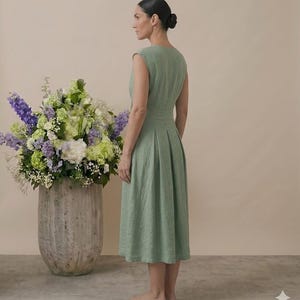 May include: A sage green sleeveless midi dress featuring a fitted bodice, a defined waistline, and a pleated skirt that falls below the knees. The garment is constructed with a simple round neckline and a clean, structured silhouette.
