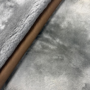 May include: A close-up shot of a grey faux fur fabric with a brown suede backing. The fur has a soft, plush texture. The suede backing is a solid, warm brown colour. The fabric is likely used for apparel or home decor.