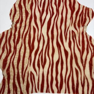 May include: A piece of fabric with a zebra stripe pattern. The stripes are a deep red color against a cream-colored background. The fabric has an irregular shape and a soft texture, suggesting it may be a hide or a similar material.