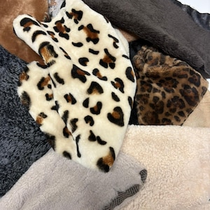 Puede incluir: Una colección de muestras de tela de piel sintética de varios colores y texturas. Incluye estampados de leopardo, telas crema, marrón y gris. Algunas muestras tienen pelo corto, mientras que otras son largas y peludas.