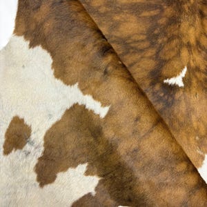 Puede incluir: Primer plano de una alfombra de piel de vaca con un patrón natural. La alfombra presenta una mezcla de manchas marrones, beige y blancas, creando una apariencia única y rústica. La textura de la piel es visible, lo que aumenta su atractivo auténtico. La alfombra está doblada, revelando la parte inferior.