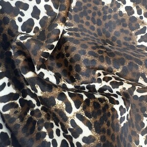 Puede incluir: Primer plano de una tela con estampado de leopardo. La tela presenta un patrón repetitivo de manchas marrones oscuras sobre un fondo beige y blanco. La tela está doblada, creando sombras y textura.