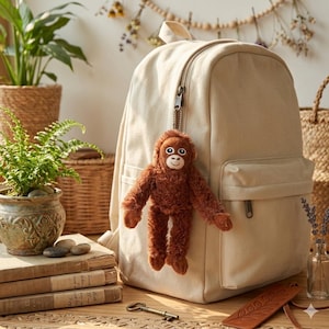 May include: A beige backpack with a front pocket and a zipper closure. A brown plush orangutan keychain hangs from the zipper. The background includes plants in pots, books, and decorative items.
