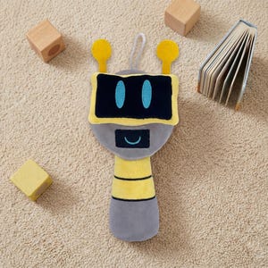 May include: A plush robot toy in gray, yellow, and black, with a smiling face and blue eyes. The toy has a loop for hanging and is surrounded by wooden blocks and a small book.