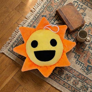 May include: A plush sun-shaped pillow with a smiling face. The pillow has a yellow center with black eyes and a large black mouth, surrounded by orange rays. A white loop is attached at the top. The pillow rests on a patterned rug.