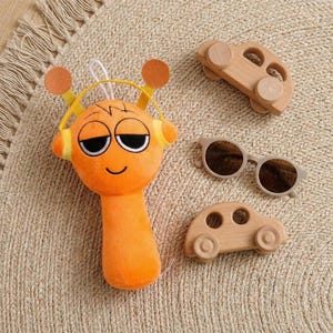 May include: An orange plush toy with a smiling face and headphones, alongside two wooden toy cars and a pair of sunglasses. The toys are arranged on a beige woven surface, creating a playful scene.