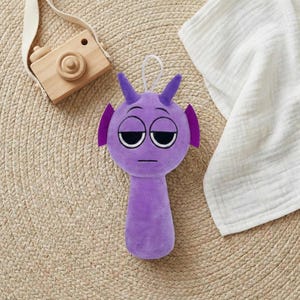 May include: A plush purple alien toy with large, sleepy eyes, small horns, and ear-like appendages. The toy has a white string for hanging and a cylindrical body. A wooden toy camera is in the background.