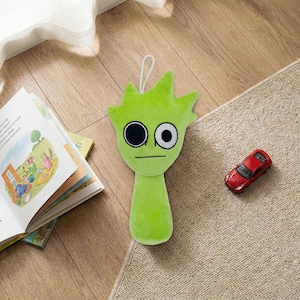May include: A lime green plush toy shaped like a cartoon character with large black and white eyes and a stern expression. The toy has a loop for hanging. A red toy car and an open children's book are also visible.