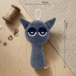 May include: A gray plush cat toy, approximately 8 inches tall and 4 inches wide, with large, sleepy eyes and pointed ears. The toy has a white loop for hanging. Other items in the image include a wooden spool and buttons.