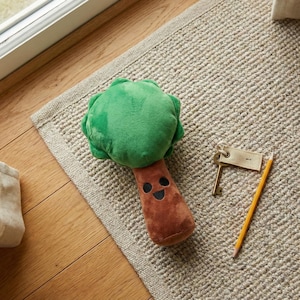 May include: A plush tree-shaped dog toy with a green top and brown trunk, featuring a smiling face. The toy rests on a beige rug, next to a pencil and keys.