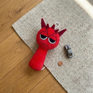 Könnte beinhalten: Ein rotes Plüschtier mit einem teuflischen Gesicht, Hörnern und einem mürrischen Ausdruck. Ein kleines silbernes Spielzeugauto und ein Cent-Stück liegen neben dem Spielzeug auf einem strukturierten beigefarbenen Teppich. Das Spielzeug liegt auf einer Holzoberfläche.