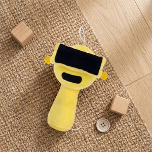 May include: A yellow plush toy with a black rectangle across the eyes, resembling a VR headset. The toy has a handle and small yellow circles on the sides. A white string is attached to the top. Wooden blocks and a button are also in the image.