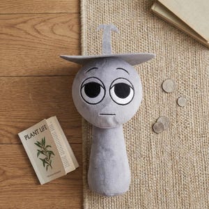 May include: A gray plush doll with large black eyes, a small mouth, and a hat-like structure. The doll is on a beige rug with a small book titled "PLANT LIFE" and some coins nearby. The doll's design is simple and cartoonish.