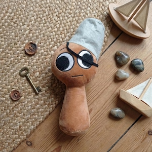 May include: A plush pirate doll with a brown body, large white eyes, and a black eye patch. The doll wears a gray hat. Wooden toy sailboats, small stones, a vintage key, and buttons are scattered around the doll.