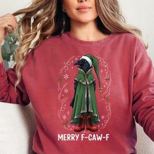 May include: Maroon sweatshirt featuring a cartoon crow dressed in a green cloak and hat, with red boots. The crow is surrounded by a festive design with snowflakes and the text "MERRY F-CAW-F" in white.