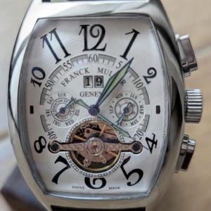 May include: A silver-toned tonneau-shaped wristwatch with a white guilloché dial, black Arabic numerals, and a brown leather strap. The face displays the text Franck Muller Geneve, a date window showing 19, two sub-dials, and an open tourbillon.
