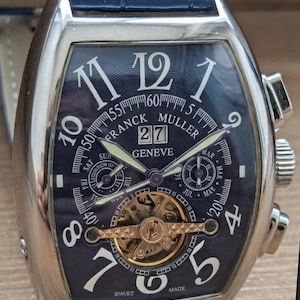 Op de afbeelding: Een Franck Muller Geneve polshorloge met een tonvormige zilveren kast en een blauwe, getextureerde wijzerplaat. Het horloge heeft grote witte Arabische cijfers, een datumvenster op 12 uur, twee sub-wijzerplaten, een gouden tourbillon en een blauwe band.
