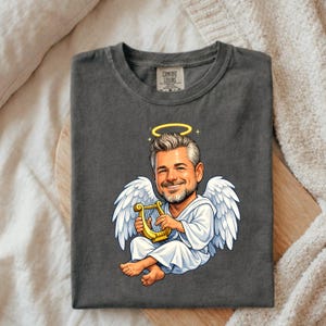 May include: Dark gray t-shirt featuring a cartoon angel with a halo, white wings, and a gold harp. The angel is smiling and wearing a white robe. The shirt has a Comfort Colors tag.