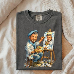 May include: Dark gray t-shirt featuring a cartoon illustration of an artist painting a portrait of himself as an angel. The artist wears a blue beret, scarf, and paint-splattered coat. A wooden easel holds the portrait. The shirt has a Comfort Colors tag.