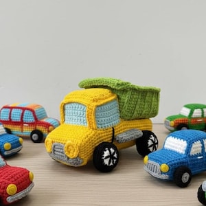 Può includere: Una collezione di auto giocattolo all'uncinetto colorate, tra cui un camion ribaltabile giallo e diverse auto più piccole in rosso, blu, verde e arancione. I giocattoli sono disposti su una superficie di legno chiaro, mostrando i loro dettagli fatti a mano.