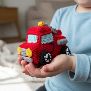 May include: A handmade red crochet tow truck toy with light blue windows, yellow headlights, and black wheels. The toy is held in a person's hands. The tow truck has gray accents and a yellow light on top.