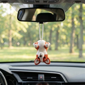 May include: A crocheted corgi ornament hanging from a rearview mirror. The small plush dog is orange and white with black eyes and a brown nose, suspended by a white cord. It is positioned upside down with its paws facing upward.