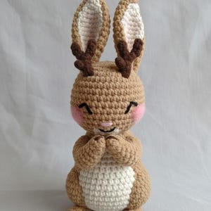 May include: A handmade crochet bunny doll in shades of brown and white, with small brown antlers and pink cheeks. The bunny has its paws clasped together and a smiling face, creating a charming and whimsical appearance. This soft toy is perfect for children.