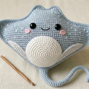 May include: A handmade crochet stingray plush toy in light blue with a white belly. The toy has black safety eyes, pink cheeks, and a smiling mouth. A wooden crochet hook rests beside the plush. The toy is approximately 12 inches wide.